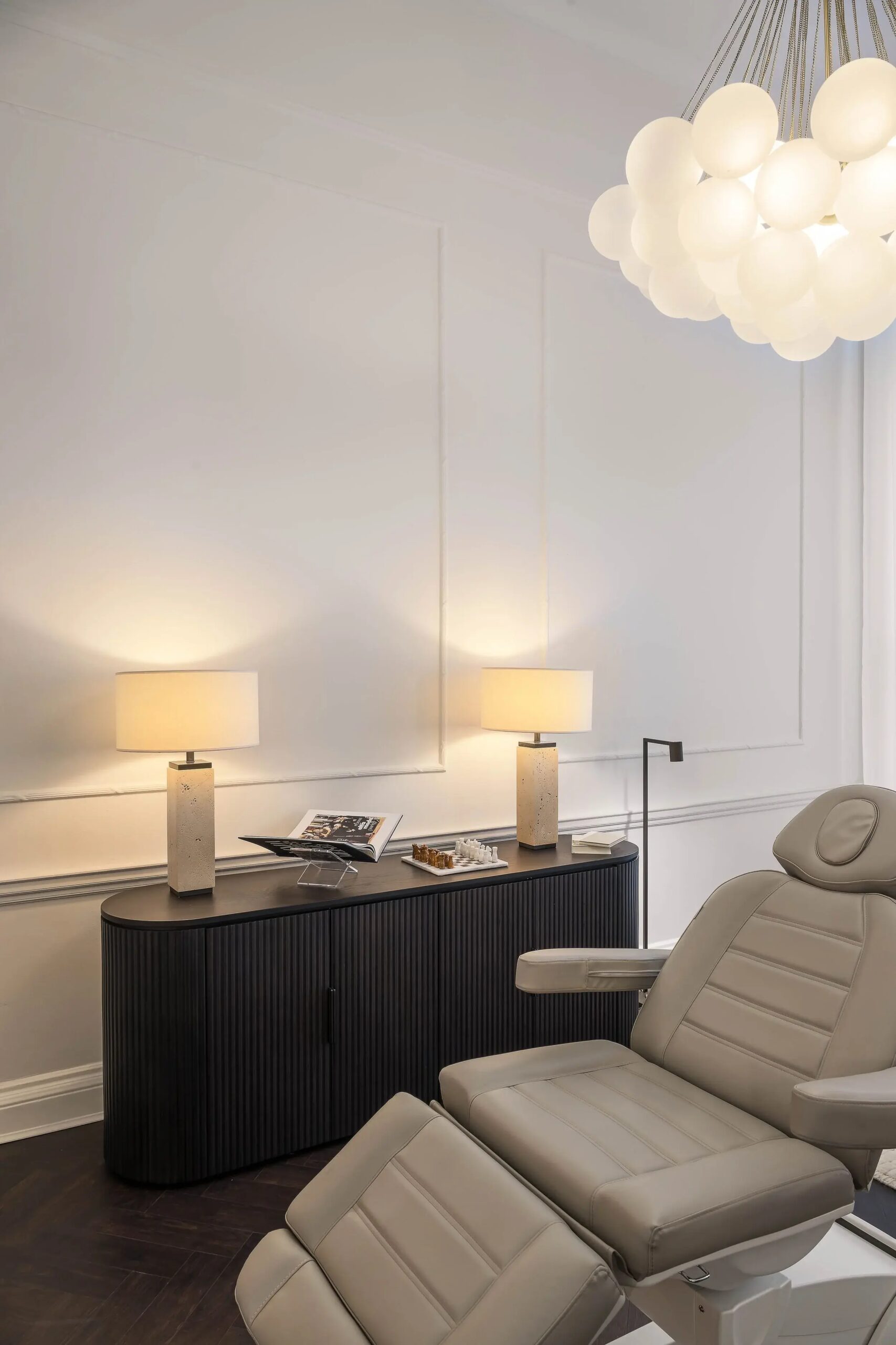 A modern dental or medical office room with a beige reclining chair, a black cabinet, two table lamps, and a ceiling light fixture with multiple globes.