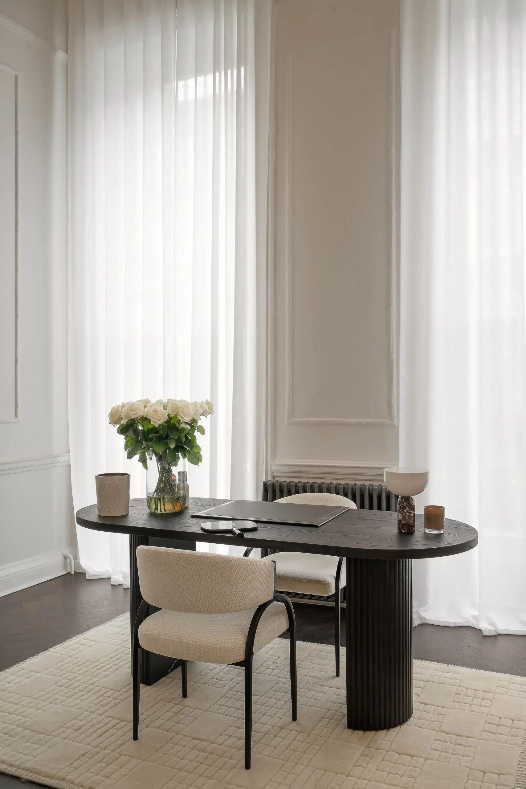 A dark wooden oval dining table with beige upholstered chairs, large white curtain panels in the background, a bouquet of white roses in a glass vase on the table, and a few decorative items including a candle, a bowl, and a closed laptop.