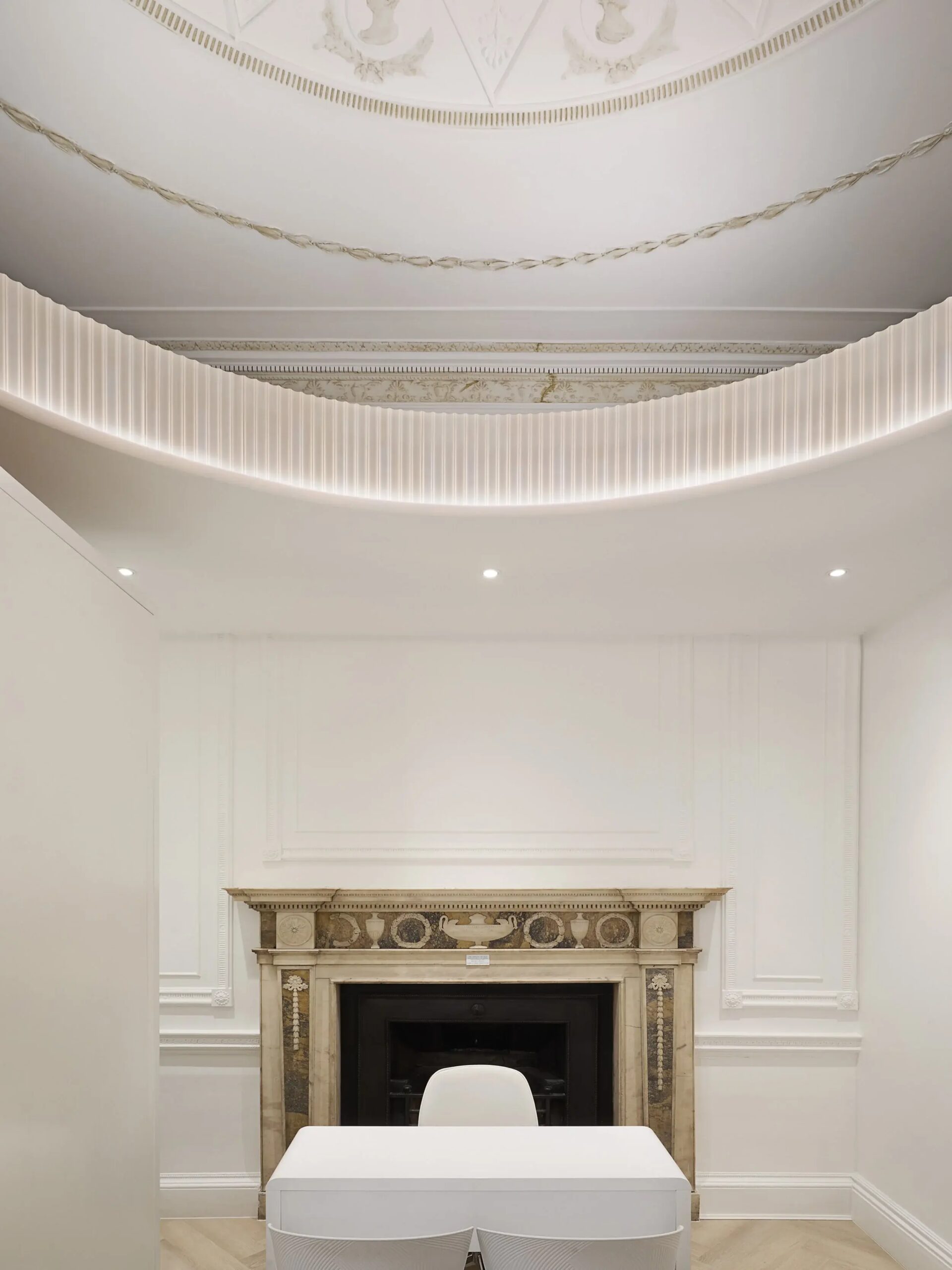 Modern interior room with white walls, a fireplace with ornate marble mantel, a white desk, white chair, and decorative ceiling moldings with lighting accents.