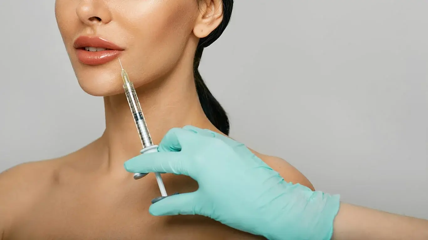 A woman receiving a cosmetic injection in her chin area from a healthcare professional wearing teal gloves.