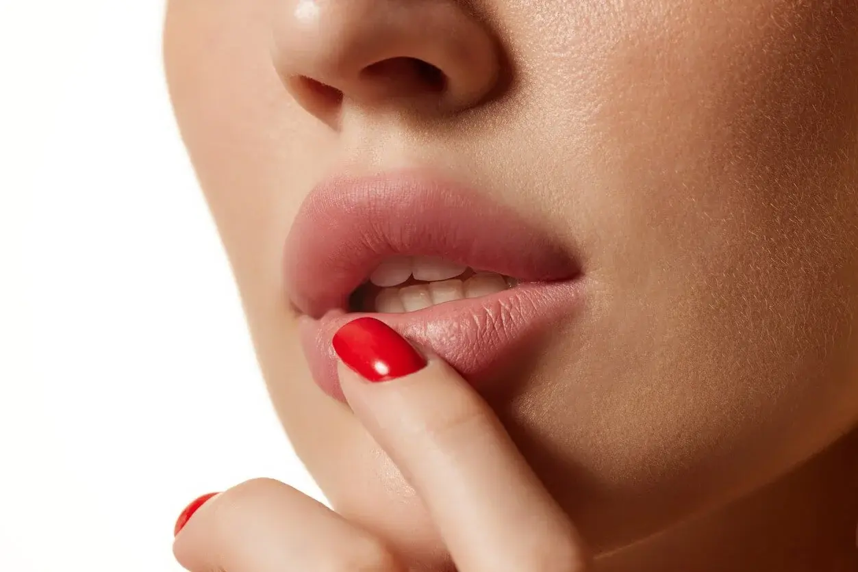 A close-up of a woman's lips with her finger touching her lower lip, lips slightly parted, with natural makeup and red nail polish.